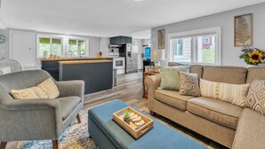 A modern living room with a grey sofa, a beige sofa, and a blue ottoman. There is a kitchen in the background with a fridge, stove, and cabinets. - Black Rapids Retreat | Waterfront Cozy Cottage (Leeds and the Thousand Islands)