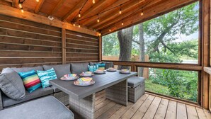 A covered outdoor seating area with a wooden deck, a table set with plates and bowls, and a view of trees through large windows. - The Riverside Retreat | Beach, Kayaks, Fire Pit (Gananoque)