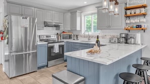 A modern kitchen with grey cabinets, stainless steel appliances, a marble island, and a coffee bar with mugs. - The Riverside Retreat | Beach, Kayaks, Fire Pit (Gananoque)