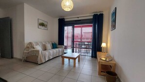 Una sala de estar con sofá, mesa de centro y mesa de comedor con sillas. Hay una ventana con persianas rojas y una puerta que da al exterior.