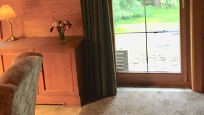 A cozy room with wooden walls, a large window with curtains, a chair, a small table with a lamp, and a painting on the wall. - Ferienwohnung im Holzhaus, 5-10 Min. vom Schönen Wandlitzsee Entfernt (Wandlitz)