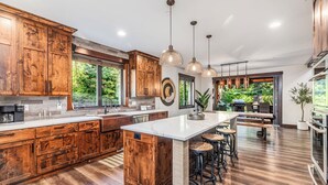 A spacious kitchen with wooden cabinets, a large island with stools, pendant lighting, and a view of greenery outside. - 1PM Check In/Out | Scenic Mountain Cabin! Private Setting+Spa/Epic GameRm (Ronald)