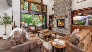 A spacious living room with leather sofas, a stone fireplace, large windows, and a television. - 1PM Check In/Out | Scenic Mountain Cabin! Private Setting+Spa/Epic GameRm (Ronald)
