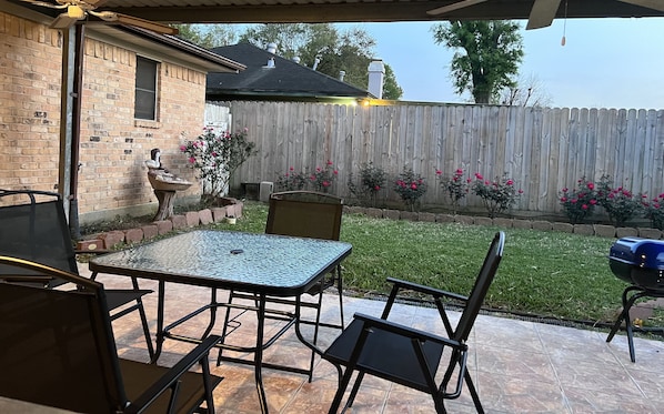 An outdoor patio with a table and chairs, a brick wall, a wooden fence, and a garden with pink flowers.