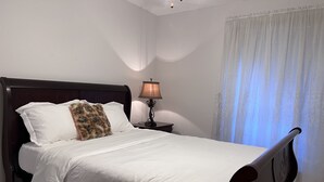 A neatly made bed with white linens, a dark wooden headboard, and a decorative throw pillow. There is a bedside table with a lamp and a curtain partially drawn to the side. - Cozy, spacious & convenient home-3bedrooms 3baths (Pasadena)
