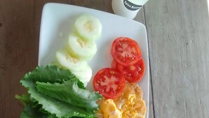 A plate with sliced cucumbers, tomatoes, lettuce, scrambled eggs, and a lemon wedge. There are two containers, one with a green lid and one with a brown lid, and a cup with a lemon slice. - Garden Guest House Bed & Bre Chitungwiza (Harare)