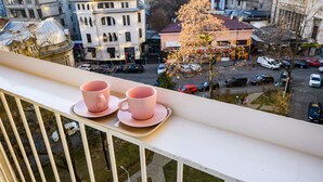 Ein Balkon mit zwei rosafarbenen Tassen auf einem Tablett, mit Blick auf eine Stadtlandschaft mit Gebäuden und einem Baum.