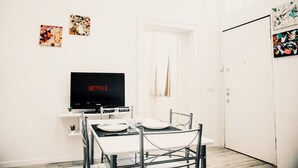 Une pièce moderne et minimaliste, dotée d’un coin repas, d’un téléviseur à écran plat affichant « NETFLIX » et d’une porte donnant sur une autre pièce.