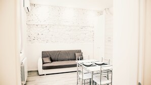 Une petite pièce moderne avec un mur en briques blanches, un canapé gris, une table blanche avec des chaises et un radiateur.