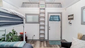 Habitación con literas, una escalera, una puerta, una ventana con persianas, una pequeña mesa con una planta y un sofá con almohadas.