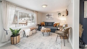 Un salón acogedor con sofá, mesa de centro y sillas de mimbre. Una ventana grande con cortinas está a la izquierda, y una lámpara a la derecha.