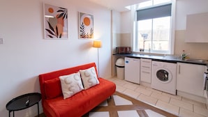 A compact living space with a red sofa, a washing machine, a small kitchen area, and a window providing natural light. - Beautiful 1-bed Apartment in Gateshead (Gateshead)