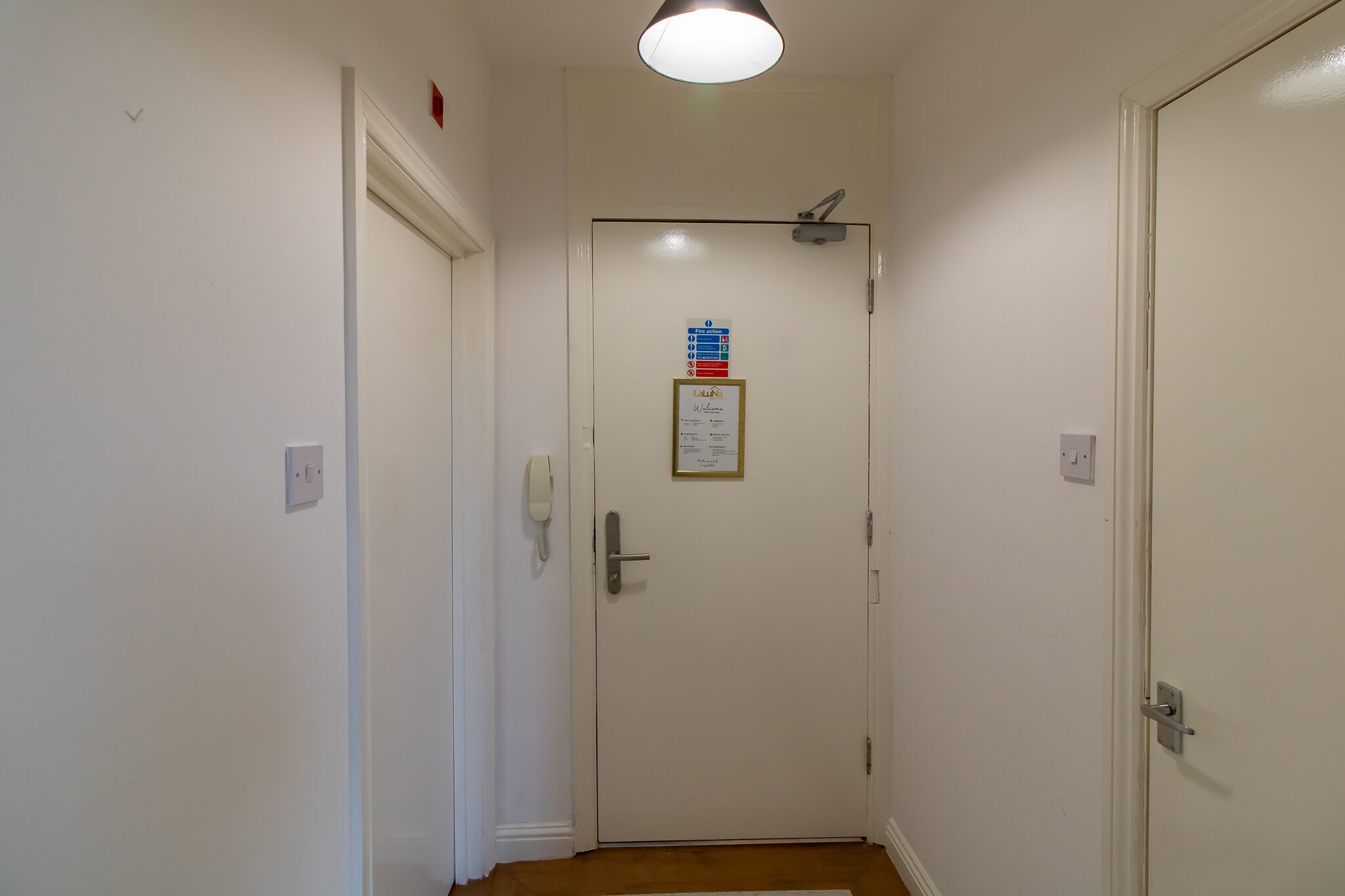 Un couloir étroit, avec une porte au fond, un luminaire au plafond et une petite pancarte sur la porte.