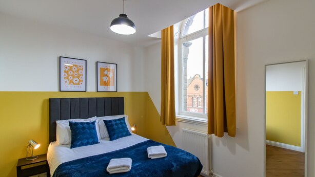 A hotel room with a bed, a nightstand, a window with curtains, and two framed pictures on the wall. - Beautiful 1-bed Apartment in Gateshead (Gateshead)