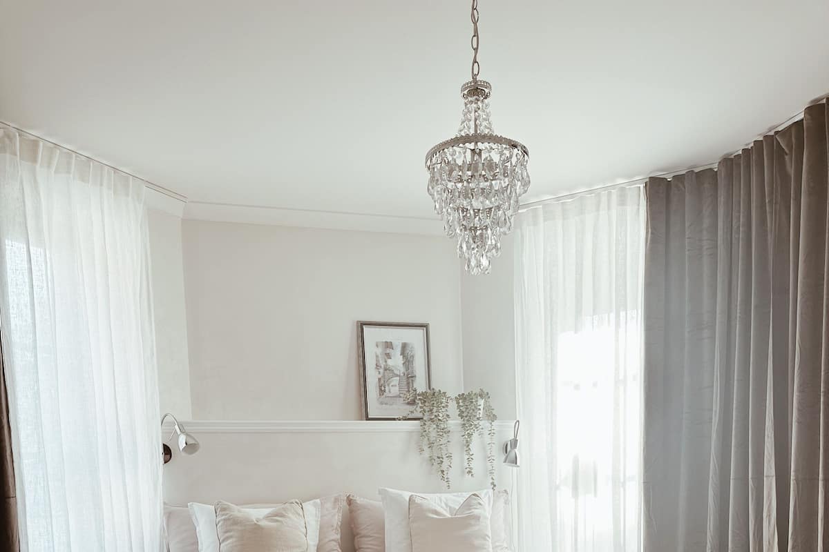 A bedroom with a large bed, a chandelier, a framed picture on the wall, and sheer curtains.
