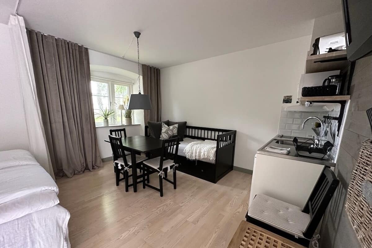 A hotel room with a bed, a dining table with chairs, a sofa, a kitchenette, and a window with curtains.