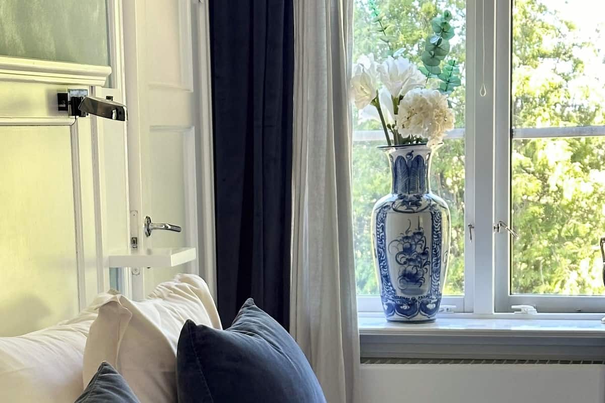 A bed with white bedding and dark pillows, a window with a vase of flowers, and a door with a handle.