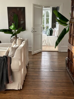 A room with a wooden floor, a white door leading to a bedroom, a large potted plant, and a cabinet. - Hallfreda Hotel (Visby)