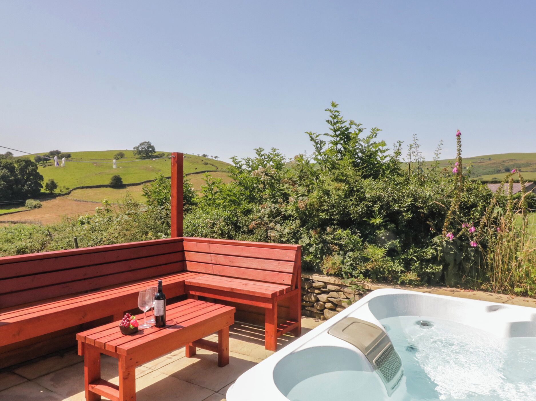 Un banc et une table en bois, un jacuzzi et une bouteille de vin, le tout installé dans un cadre verdoyant avec des collines en arrière-plan.