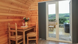 Interior de una cabaña de madera con una mesa de comedor y sillas, un florero con flores, y vistas al exterior a través de un ventanal.