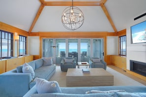 A spacious living room with a large sofa, two armchairs, a coffee table, and a chandelier. There are large windows with a view of the ocean.