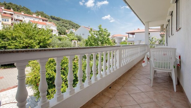 Un balcon doté d’une balustrade blanche, d’une chaise en plastique blanche et offrant une vue sur des bâtiments et de la verdure en arrière-plan.