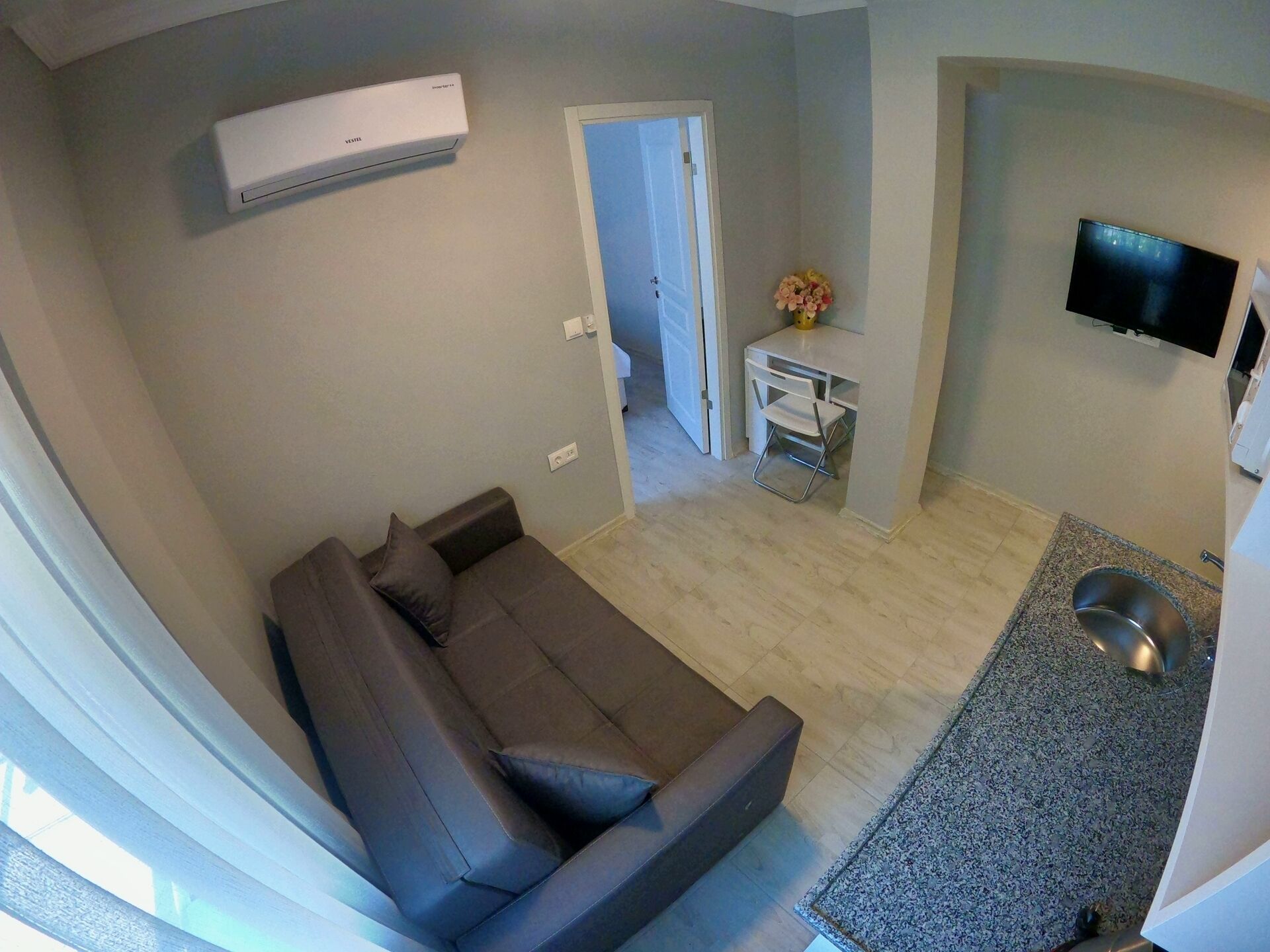 A compact living space with a sofa, a small table with a flower vase, a TV mounted on the wall, and a kitchen area with a sink and a metal bowl.