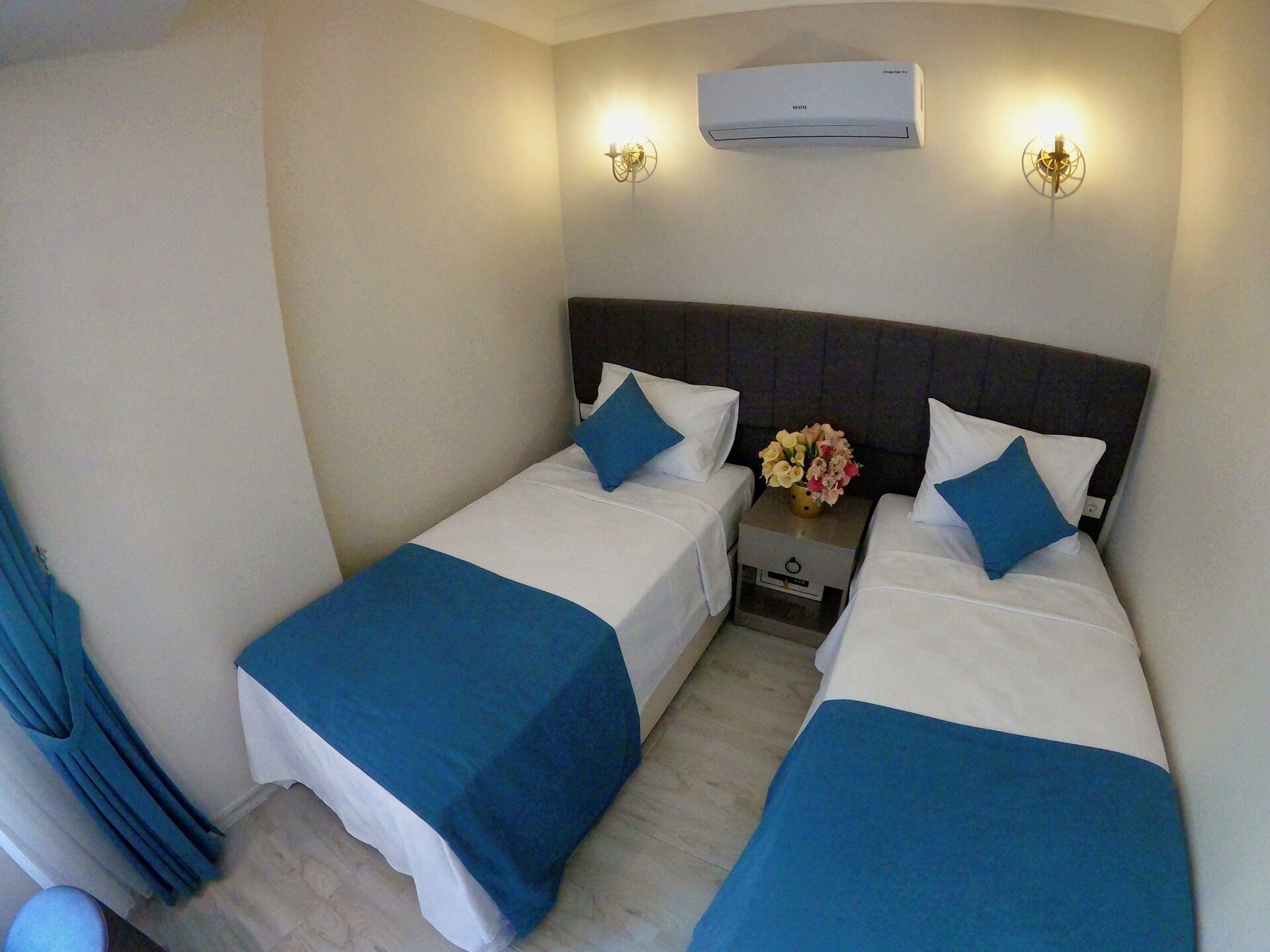 A hotel room with two beds, a nightstand with a vase of flowers, and an air conditioning unit on the wall.