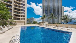 A swimming pool with lounge chairs, surrounded by tall buildings and palm trees.
