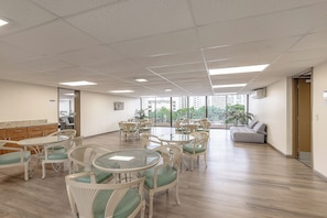 Um quarto amplo com várias mesas redondas e cadeiras, um sofá e vista para edifícios altos através de grandes janelas.
