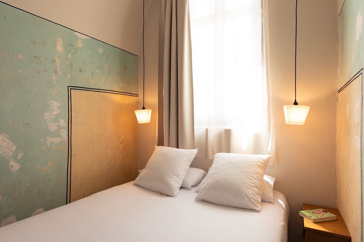 A bed with white linens and pillows, a bedside table with a book, and a window with curtains.