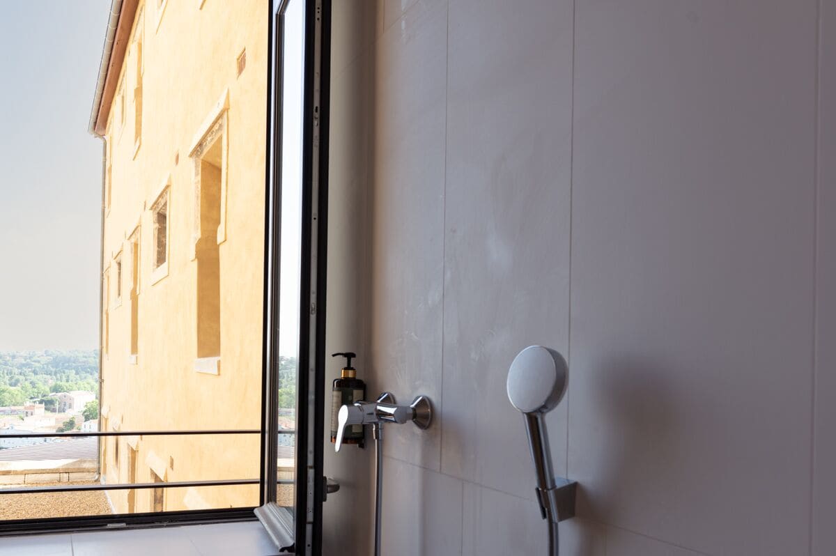 A bathroom with a shower, a view of a building outside, and a bottle of shampoo on the counter.