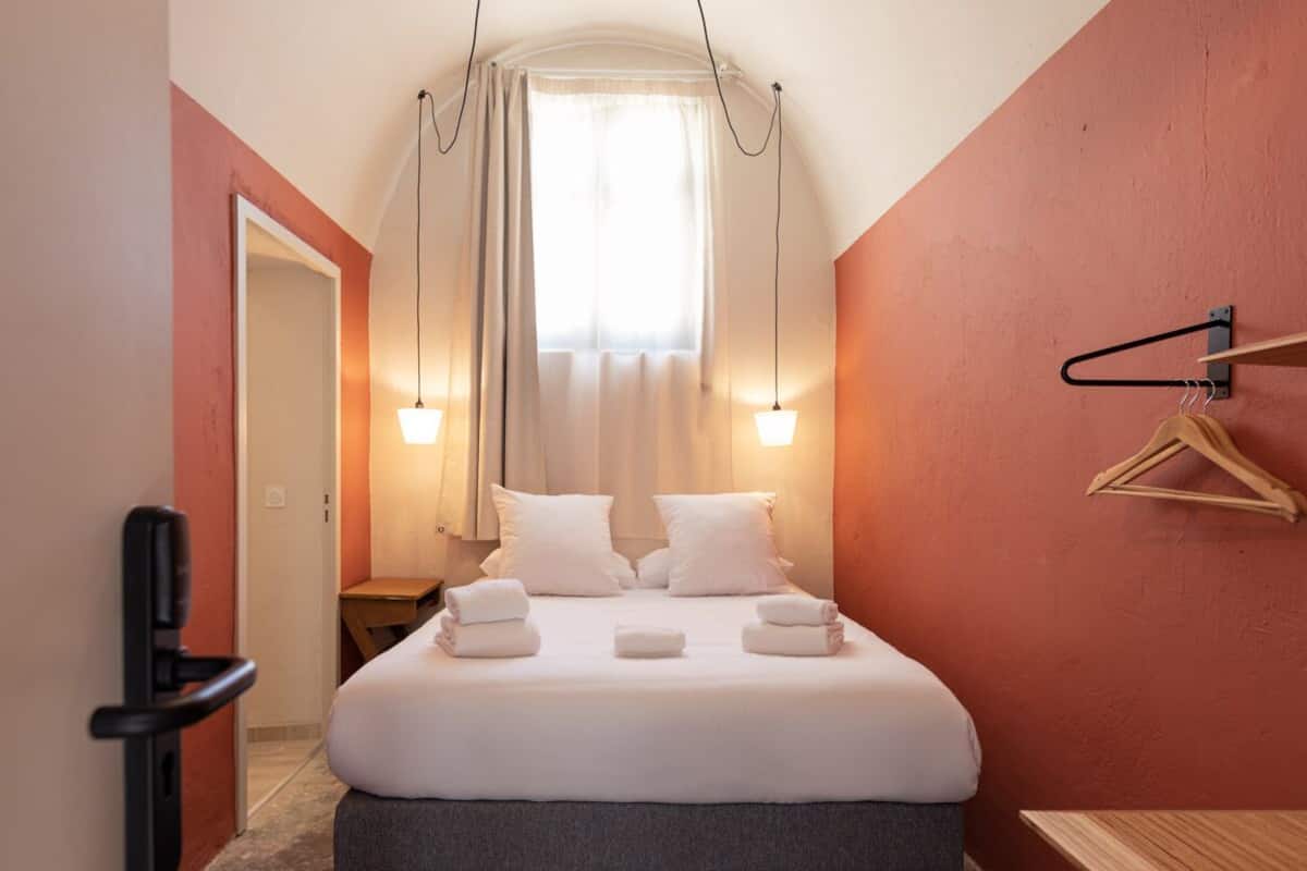 A hotel room with a bed, bedside tables, a hanging light fixture, and a coat rack.