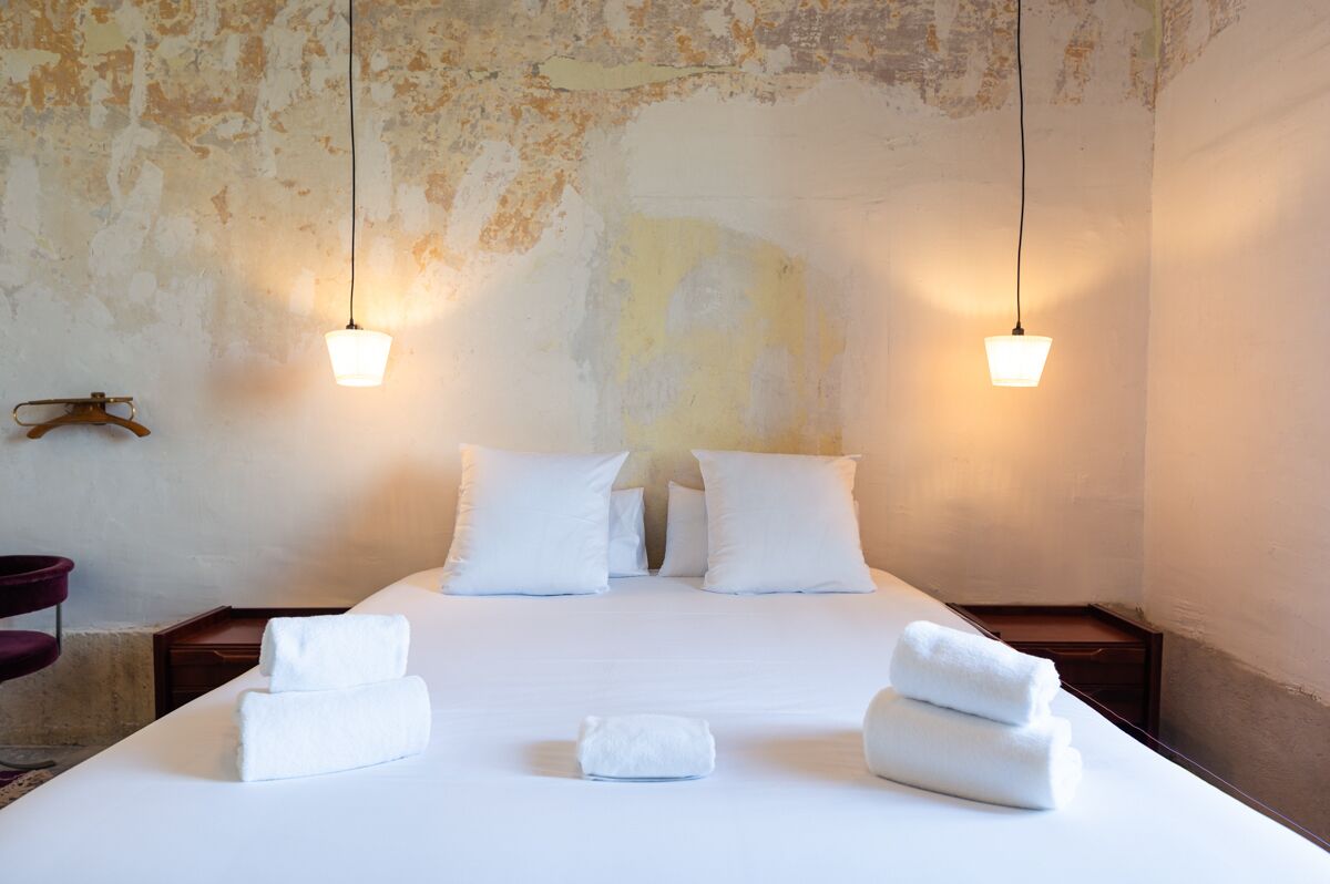 Een opgemaakt bed met witte lakens en handdoeken, twee nachtkastjes en een rustieke muur met afbladderende verf.