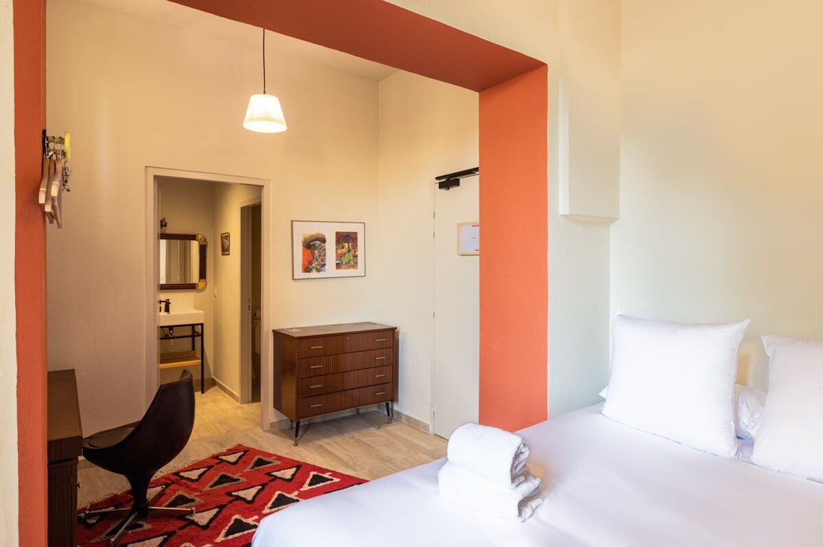 A hotel room with a bed, a dresser, a chair, a red patterned rug, and a hallway leading to a bathroom.