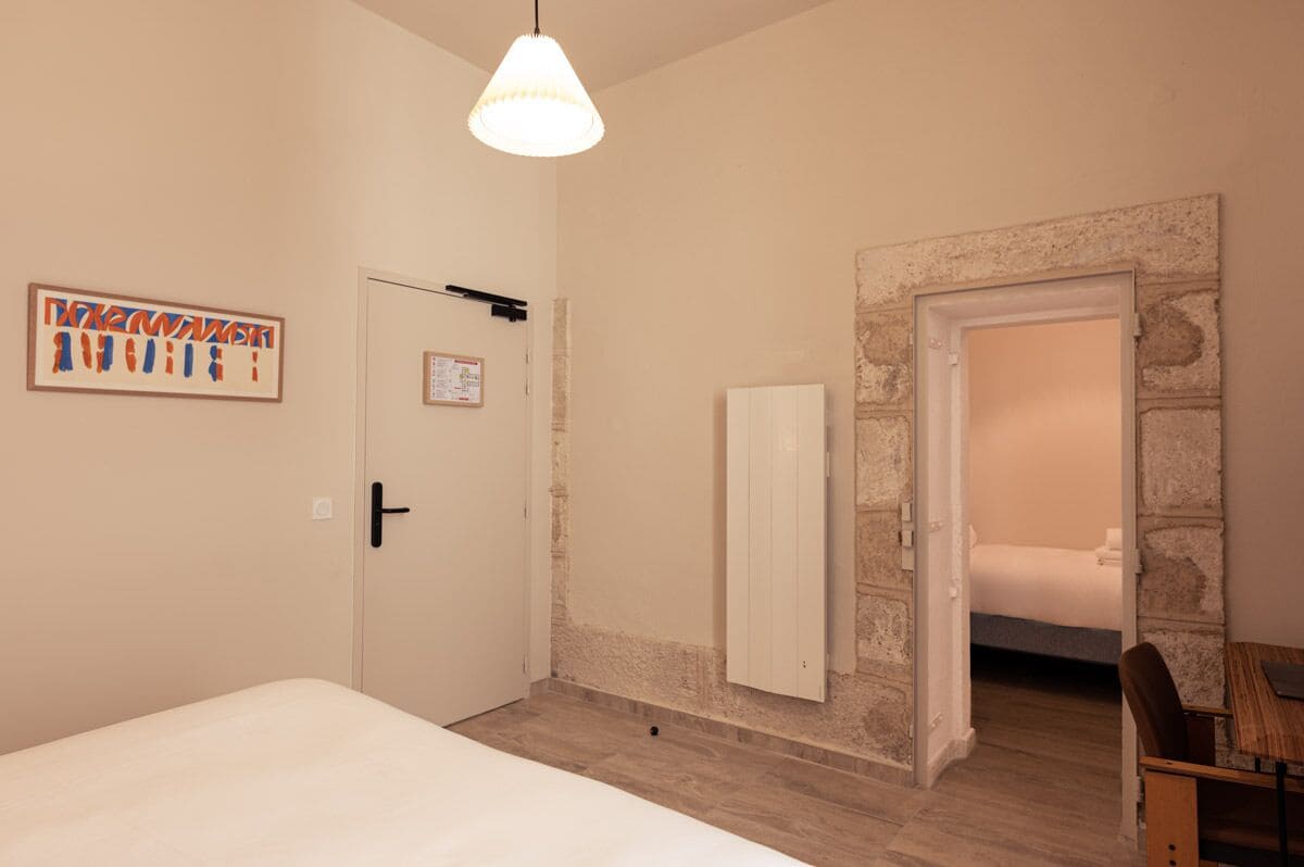 A bedroom with a bed, a desk, and a chair. There is a door with a calendar on it, and a wall with a stone accent.