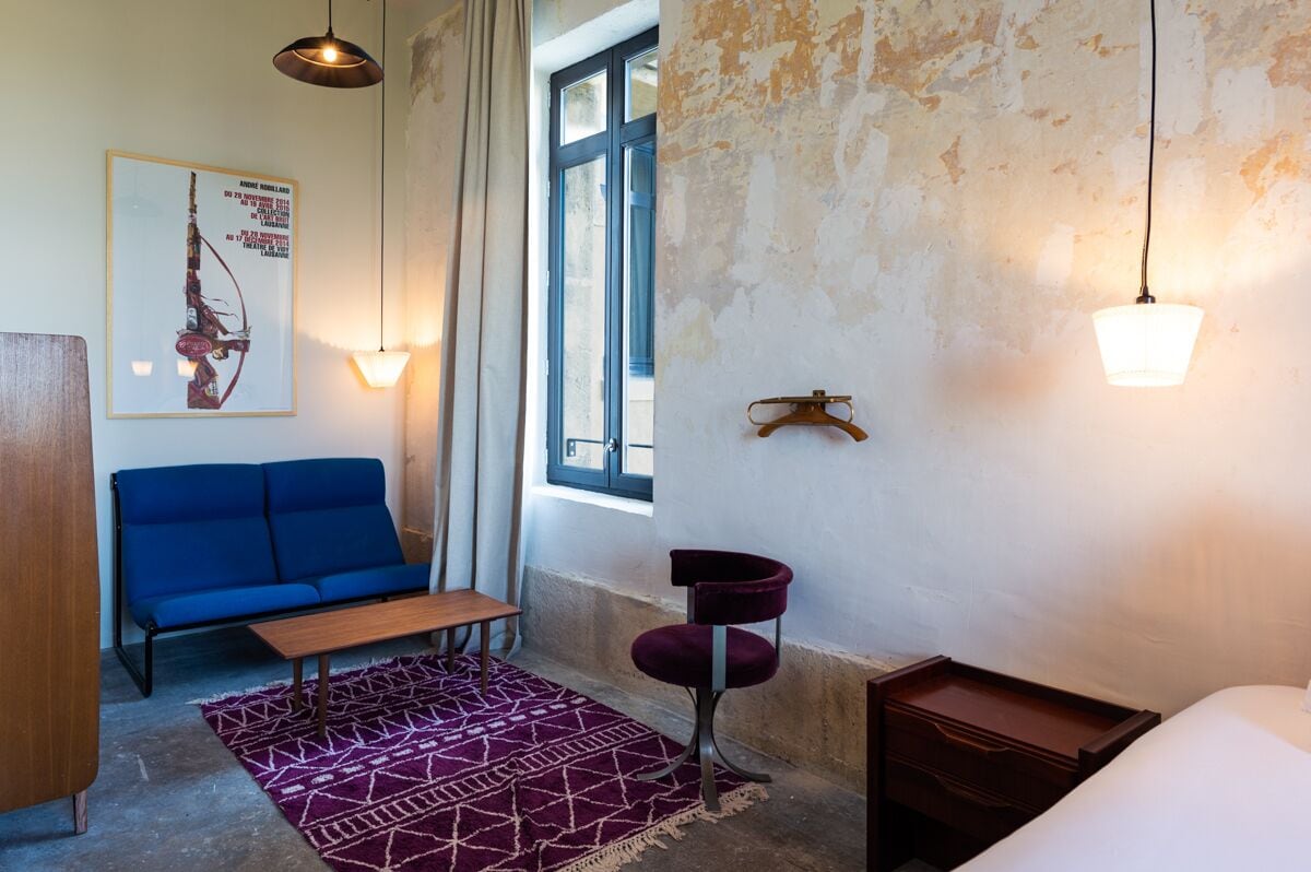 A room with a blue sofa, a wooden table, a purple chair, a bed, a hanging light fixture, and a window with curtains.