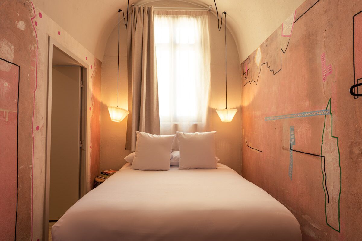 Une chambre d’hôtel étroite, avec un lit, deux lampes de chevet et une fenêtre ornée de rideaux légers.