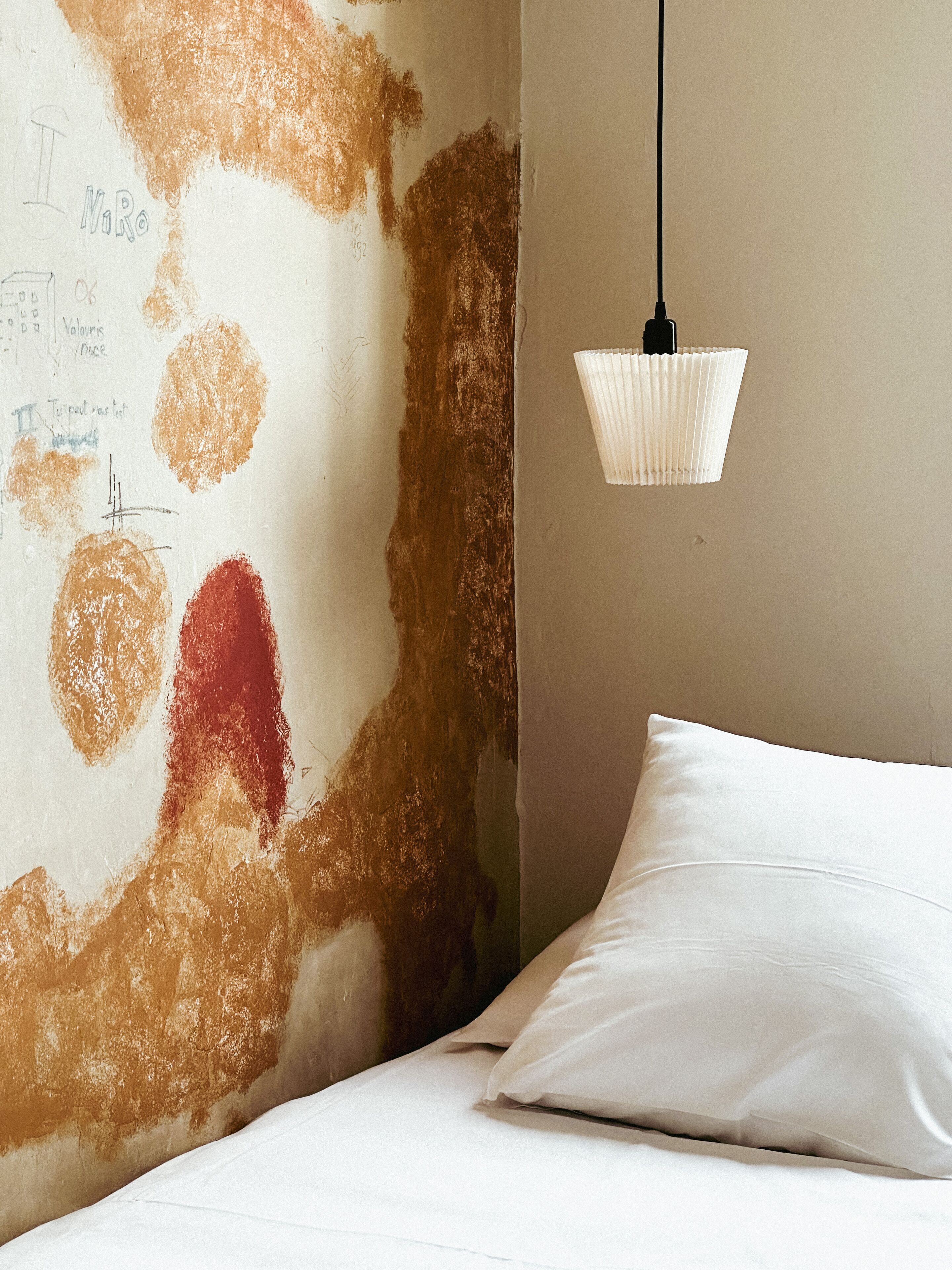 A bedroom with a bed, a white pillow, a textured wall with scribbles, and a hanging light fixture.