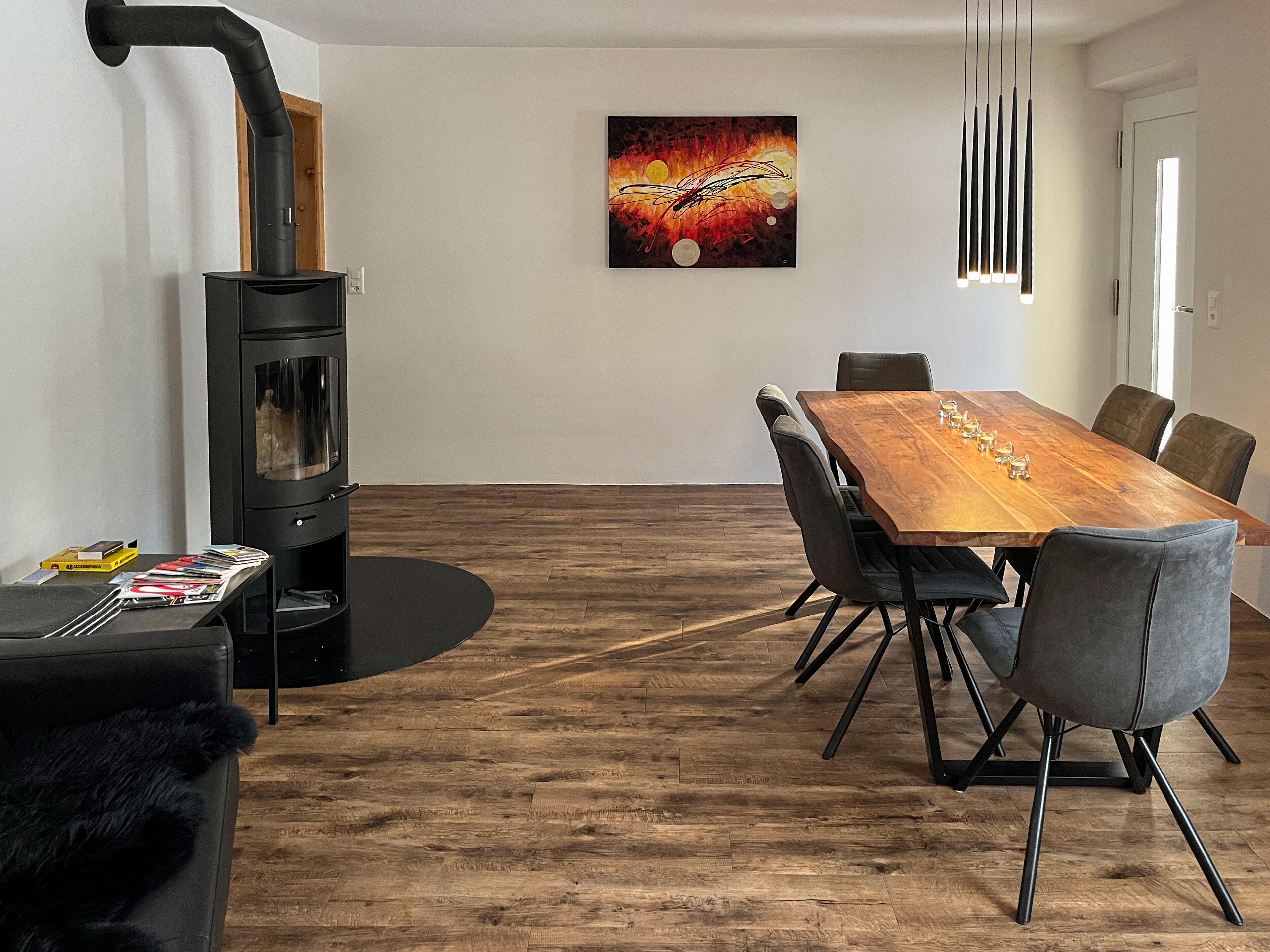 Un salon moderne avec une grande table à manger en bois, des chaises à revêtement gris, un canapé noir et un poêle à bois.