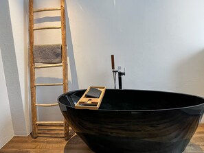 A black bathtub with a wooden tray and a smartphone on it, a wooden ladder, and a towel rack against a white wall. - Beautiful, sunny ground floor apartment with 3 rooms and garden (Poschiavo)