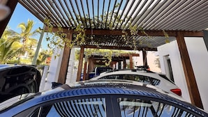 Une voiture est garée sous une pergola en bois, avec vue sur des palmiers et un bâtiment moderne en arrière-plan.