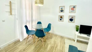 Een moderne woonkamer met een ronde glazen tafel, blauwe stoelen, een televisie op een dressoir en abstracte kunst aan de wand.