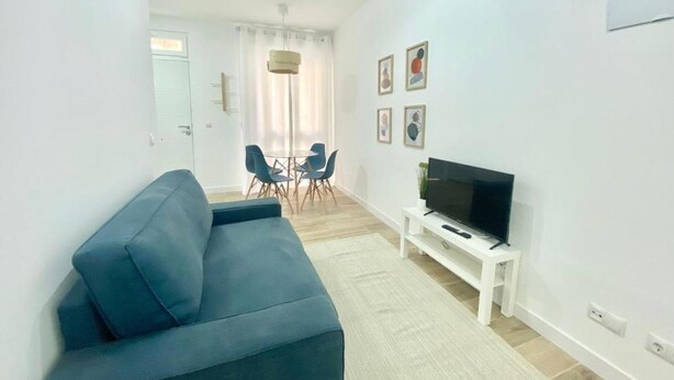 Een woonkamer met een bank, een televisie op een meubelstuk, een eethoek met stoelen en een deur.