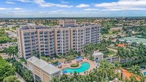 A multi-story hotel with a central pool surrounded by palm trees and lounge areas. - Elaborate Beachfront Condo | Rooftop Pool/Spa (Marco Island)