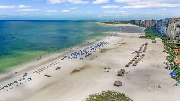 A beach with clear blue waters, a sandy shore, and a row of buildings along the coastline. - Elaborate Beachfront Condo | Rooftop Pool/Spa (Marco Island)