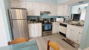 A compact kitchen with white cabinets, stainless steel appliances, and a small dining area with a blue table and wooden chairs. - One block to the beach!  5 Rose Garden (Lewes)