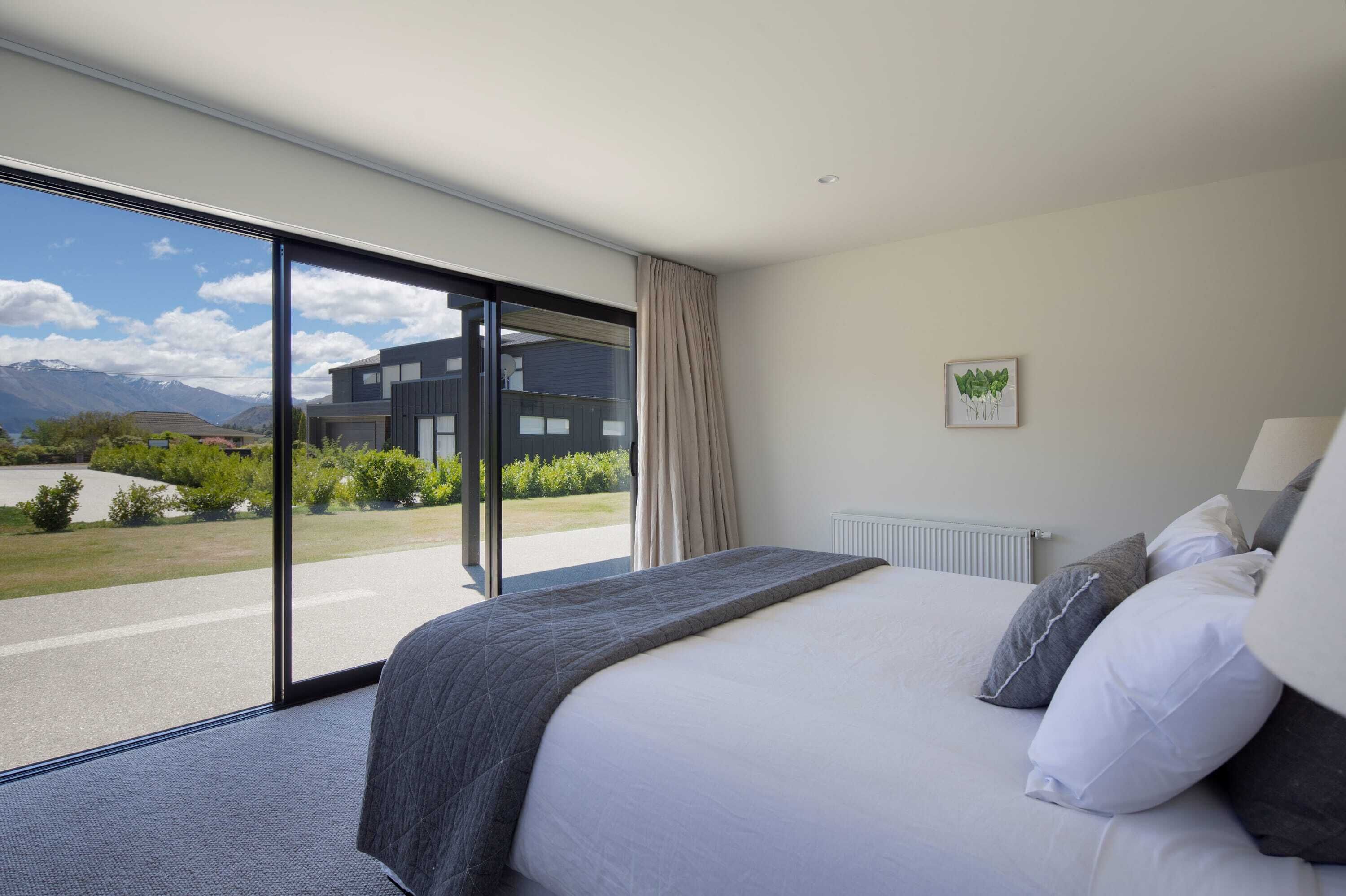 Une chambre d’hôtel moderne avec un grand lit, une vue sur un immeuble et des montagnes à travers une porte-fenêtre coulissante, et un radiateur au mur.