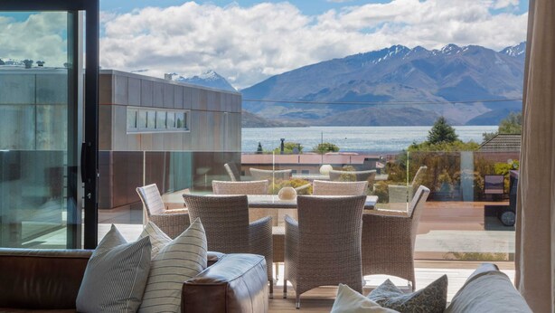 A modern interior with a glass wall, a brown leather sofa, wicker chairs, and a view of mountains and a lake. - Waimaori Views The Sp (Wānaka)
