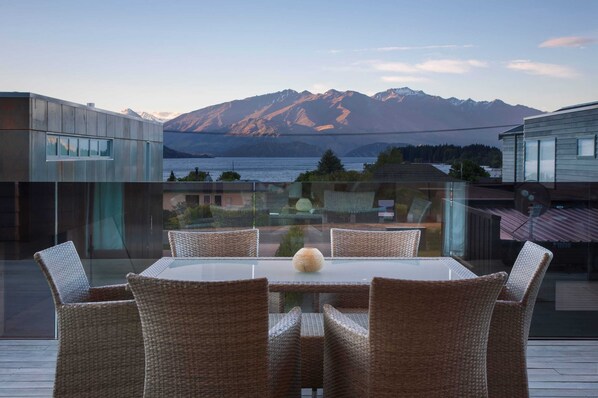 Un comedor con paredes de vidrio, sillas de mimbre y una mesa central, con vistas a un paisaje montañoso y una masa de agua.